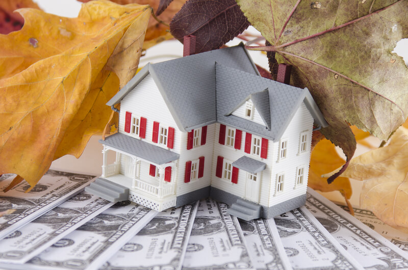 a home sits on top of dollar bills with fall leaves in the background